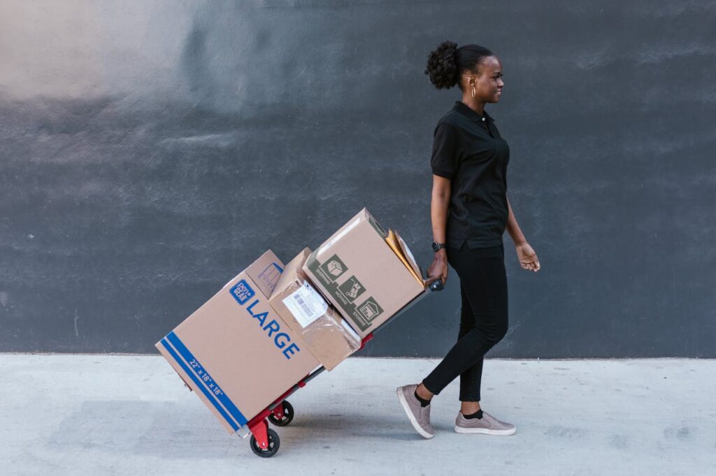 A female courier in uniform delivering packages on a trolley outdoors against a dark wall.